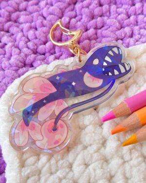 Gold keychain attached to a clear acrylic charm with a pink/purple butterfly design, resting on a purple fuzzy rug with colorful pencils nearby.