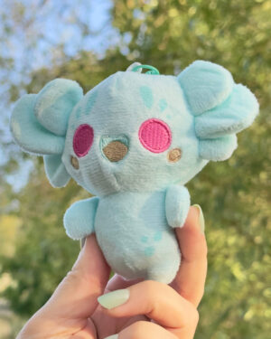 Light blue plush toy with pink cheek spots being held up outdoors, background of green trees blurred