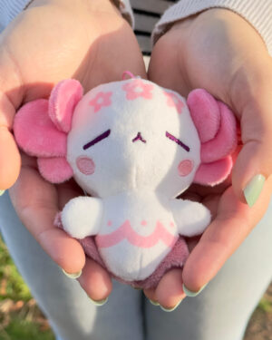 Small plush toy held in two hands; white body with pink ears, flowers, and cheeks, cute and cuddly.