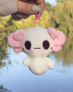 White plush toy with pink ears hangs from a pink clip, held by a hand with pale green nails, outdoors by a lake.