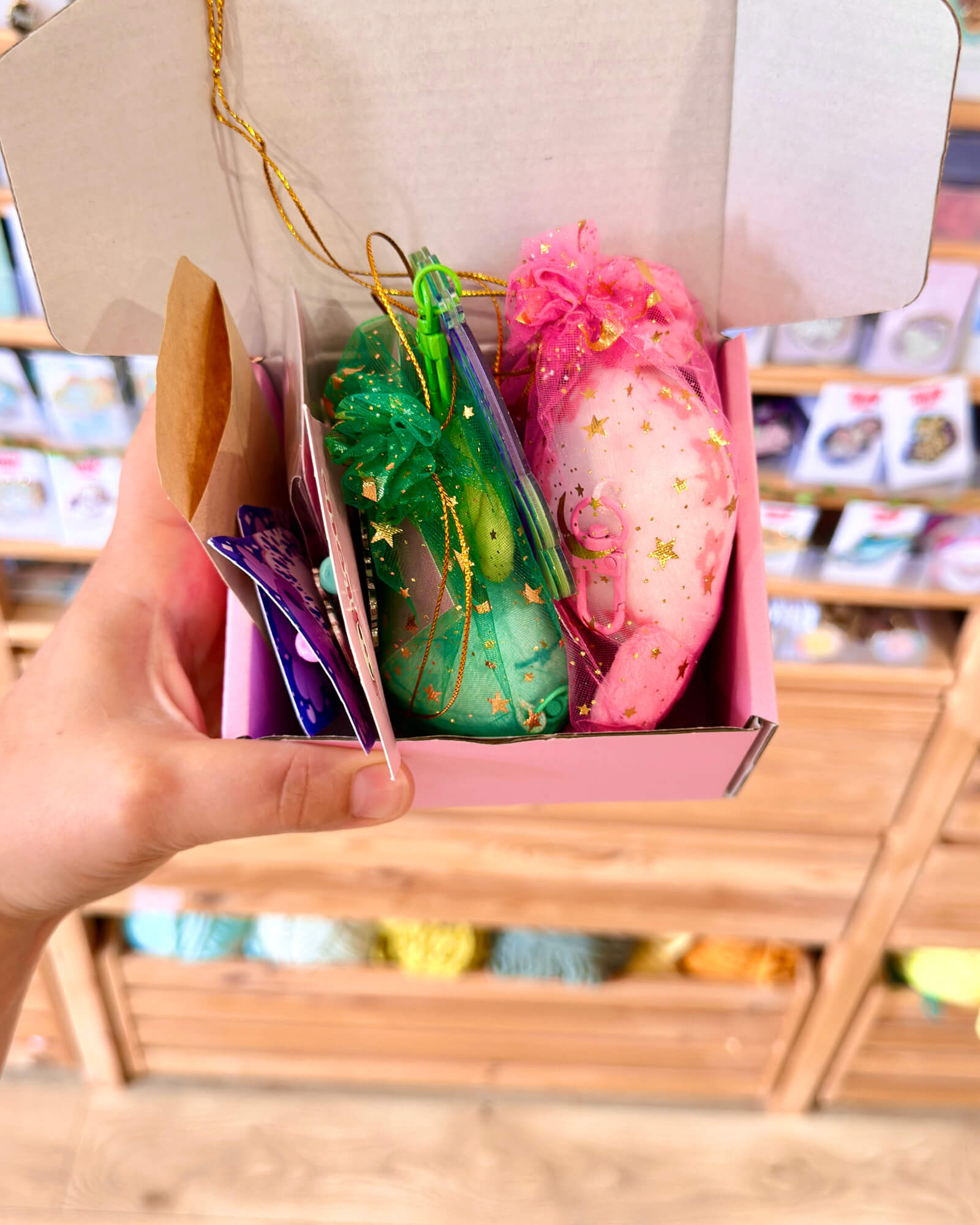 Mini Lotl Box Mystery B Grades by Evy Benita 1 - Evy Benita A hand holding a pink gift box filled with colorful organza bags decorated with gold stars in a store setting
