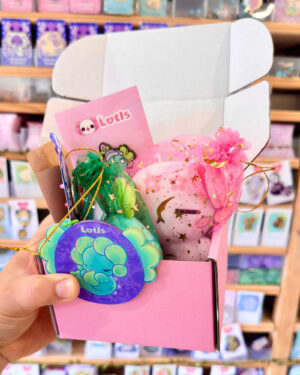 A person holds a pink gift box with decorative pink and green organza bags and Lotts cards in a toy store backdrop.