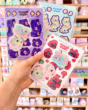 Two colorful sticker sheets with cute blob animals and desserts held up in a store display.