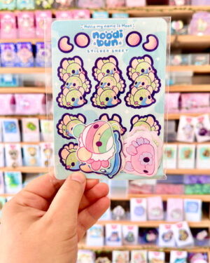Noodibun Mystery B Grade Sticker Bags B - Evy Benita Sticker sheet featuring pastel cow-like doodles and text 'Hello my name is Moos' on a blue background in a store display