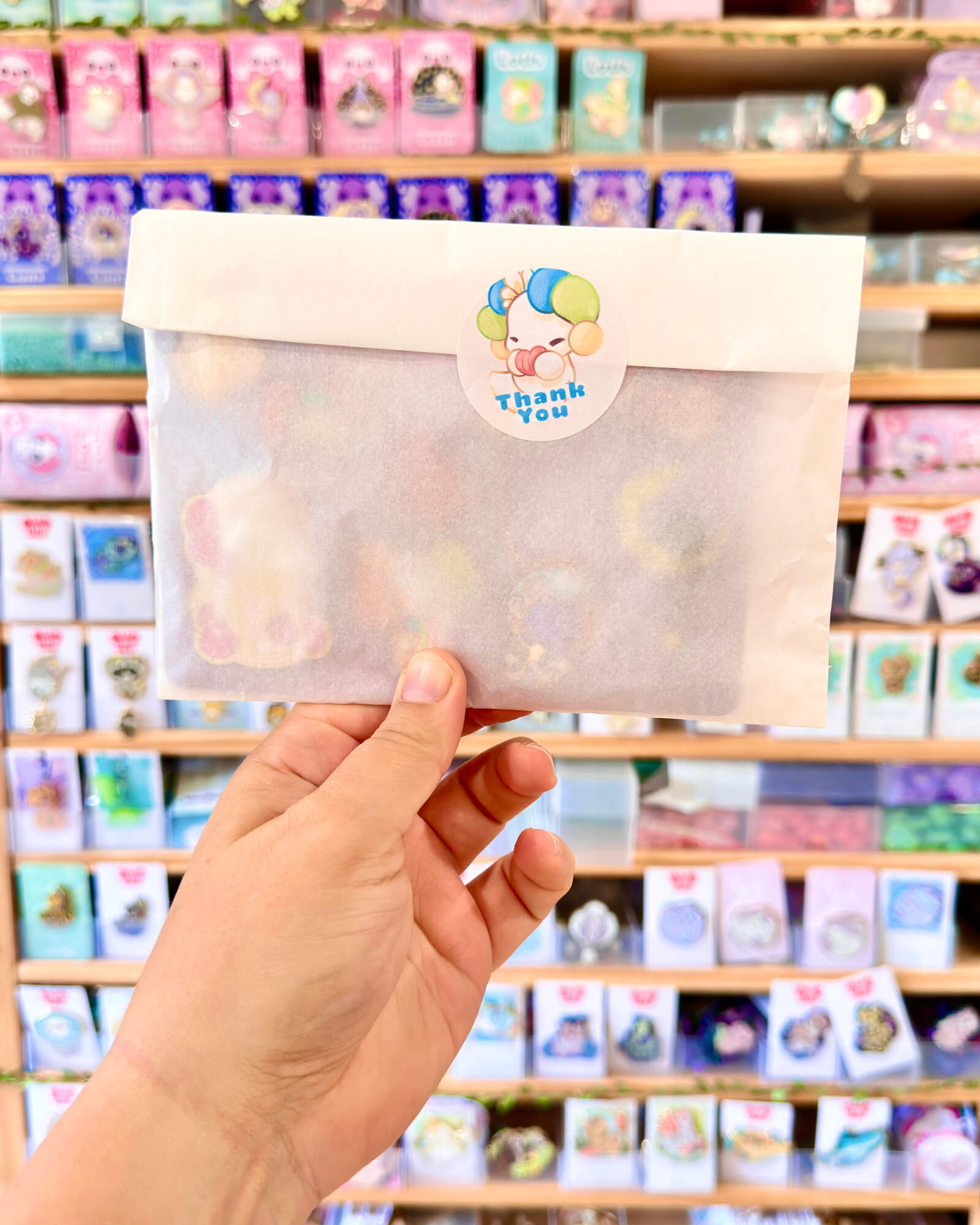 The Lotls B Grade Mystery Enamel Pin Sets 1 - Evy Benita Hand holding a white gift bag with a circular 'Thank you' sticker showing a colorful cartoon character, in a store aisle.