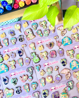 The Lotls B Grade Mystery Enamel Pin Sets 2 - Evy Benita Assorted colorful cartoon pins and keychains laid out on purple display cards labeled 'Lotis'