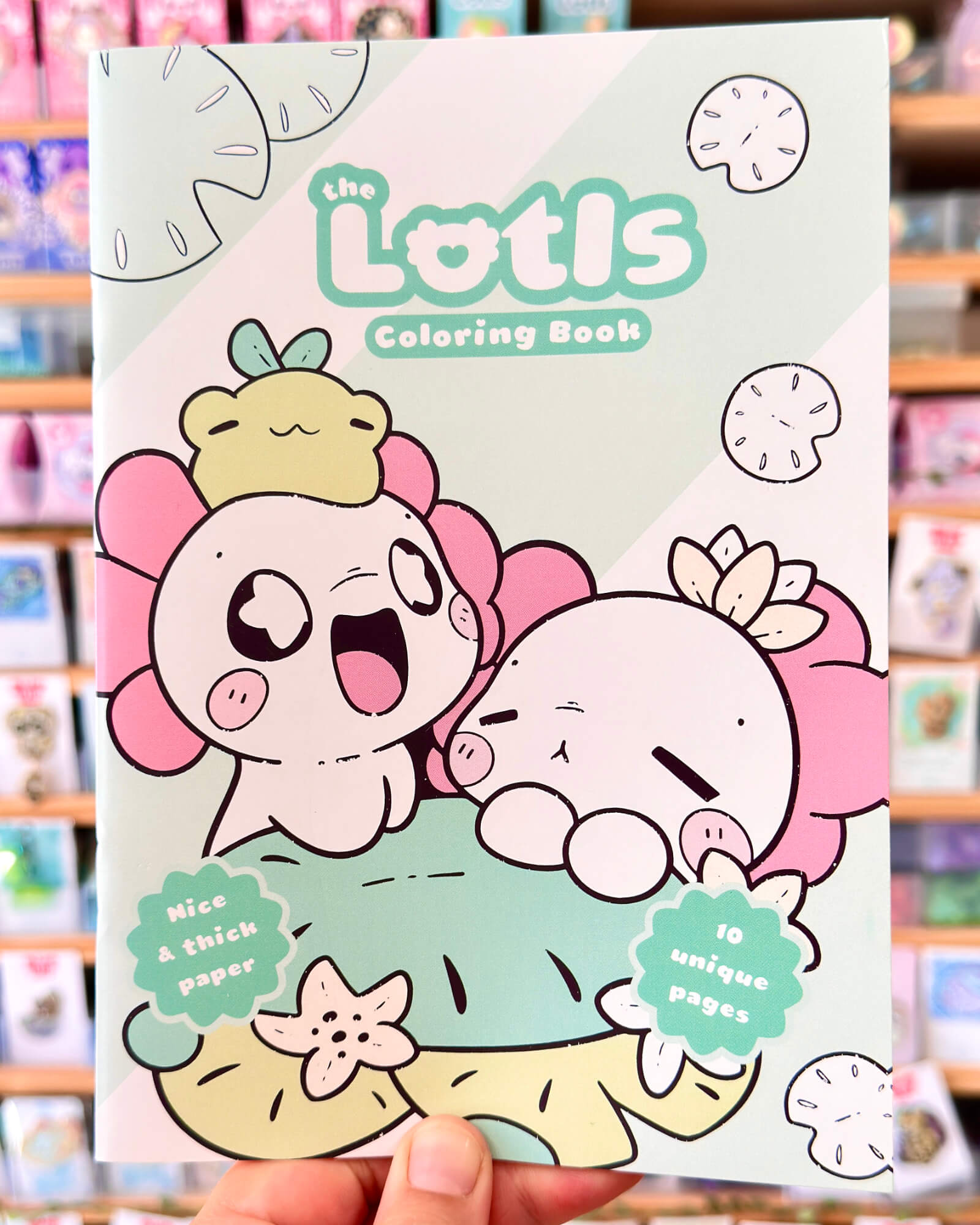 The Lotls in Lily Pond Valley Coloring Book 2 - Evy Benita Front cover of The Lottis Coloring Book with cute pink characters and pastel background, held by a hand