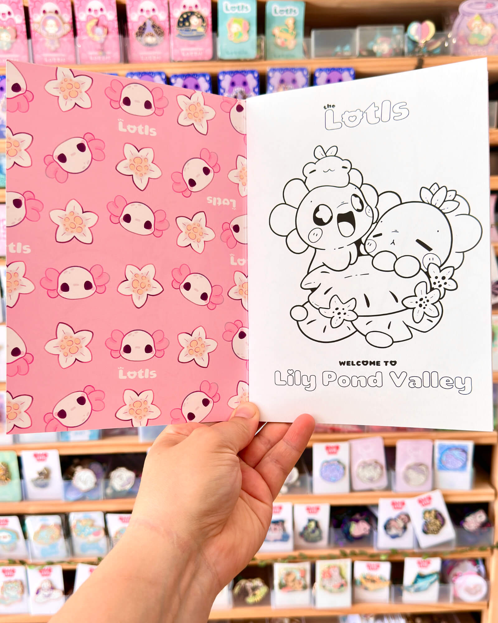 The Lotls in Lily Pond Valley Coloring Book 3 - Evy Benita Two open children's books held in a store: left page is pink with flower patterns and small character faces, right page shows a black-and-white line drawing of cuddly animals with the title Lily Pond Valley.