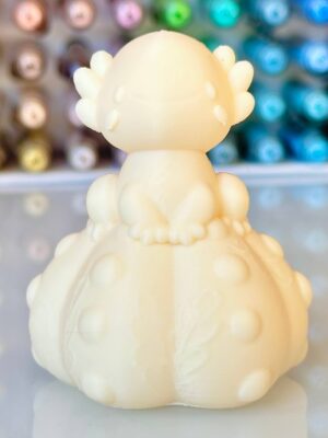 Back view of a small cream-colored plastic figurine with rounded head and ribbed collar, wings on the sides of its head, blurred colorful background
