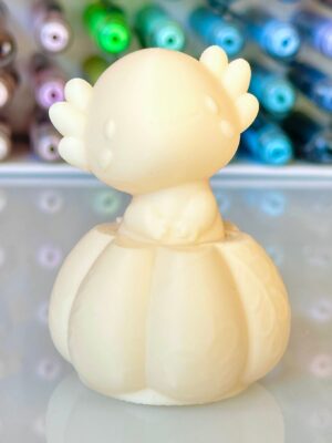Cream-colored toy dinosaur viewed from behind, with rounded body and small plate-like spikes along its back, on a reflective surface with colorful thread spools in the background.