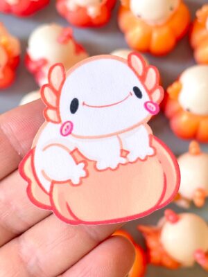 Close-up of a pink cartoon crab charm with a smiling face, being held between fingers; orange and white beads blurred in the background.